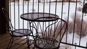 A wooden deck with a metal table and two chairs, overlooking a snowy landscape with houses and trees.