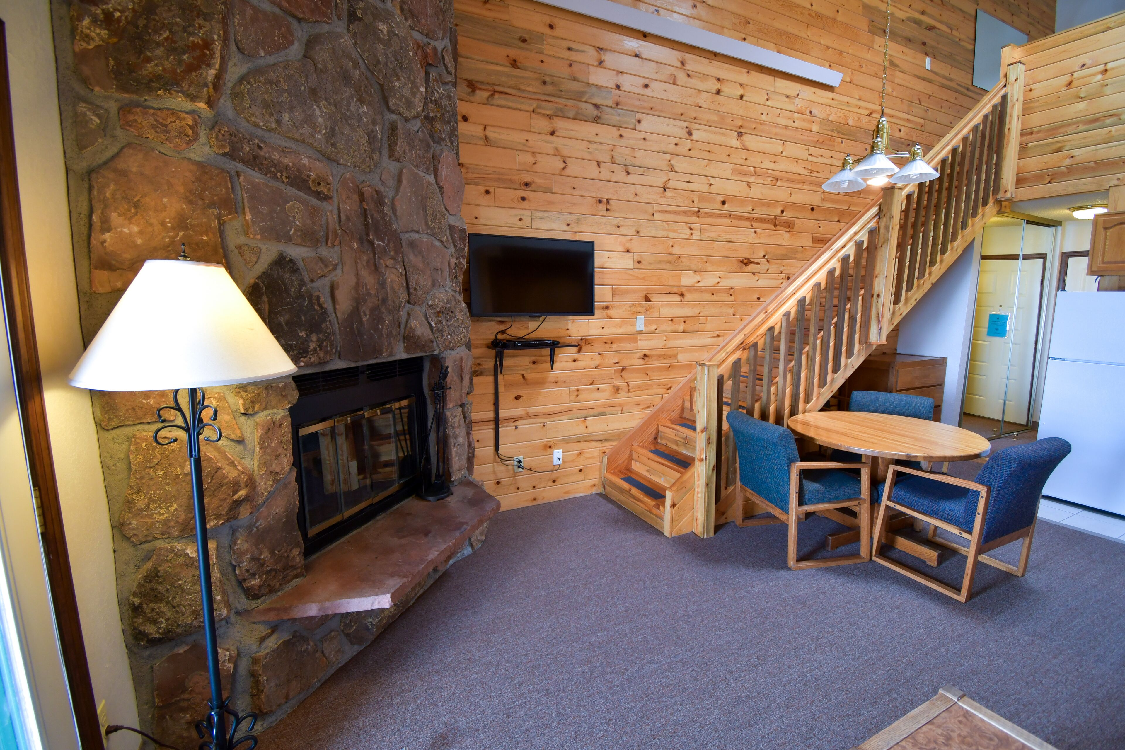 A cozy interior with a stone fireplace, wooden walls, a TV mounted above it, a staircase, and a small dining area with blue chairs.