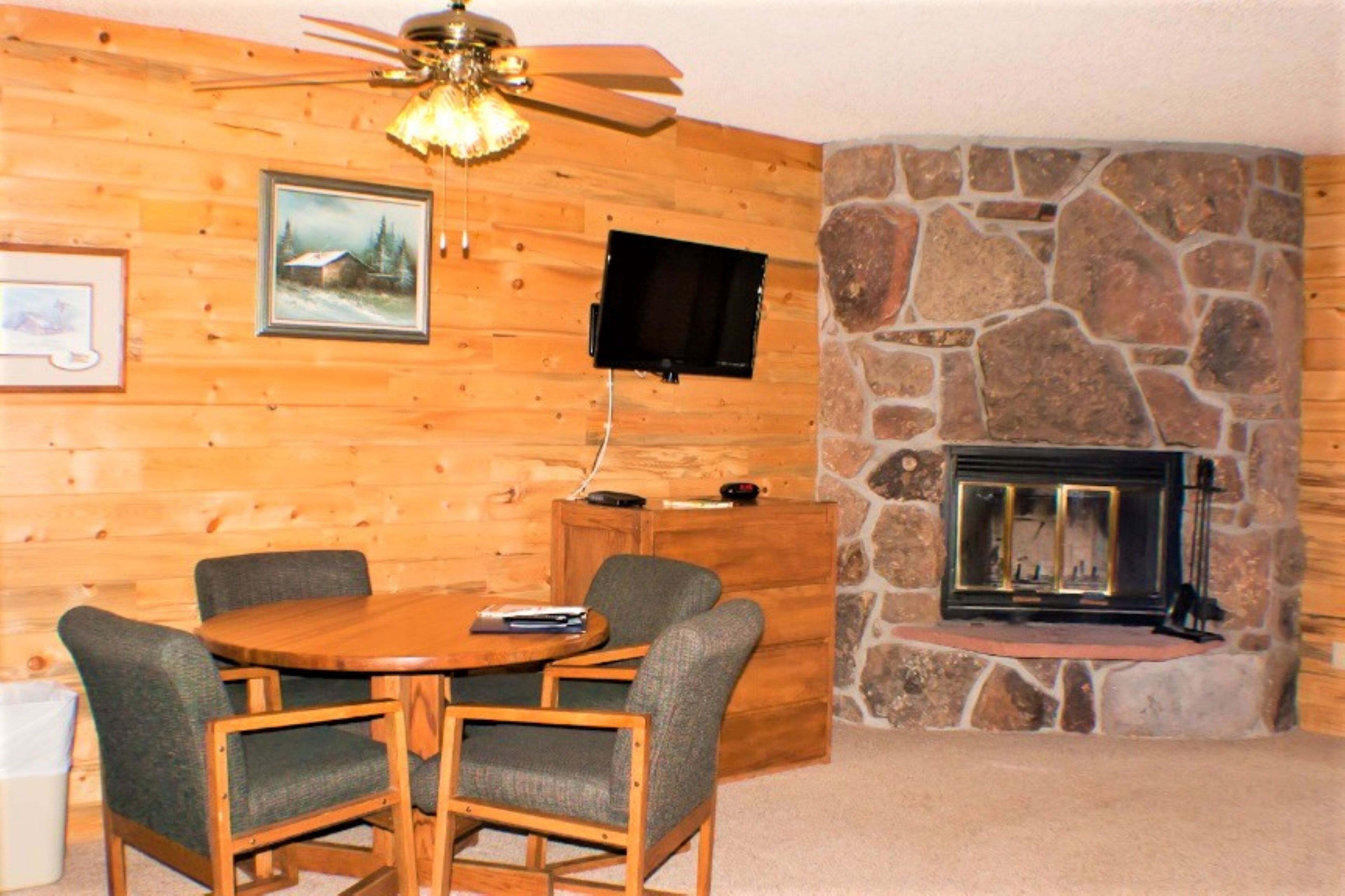 Una habitación acogedora con una pared de madera, una chimenea de piedra, un televisor montado en la pared y una mesa redonda de madera con cuatro sillas.