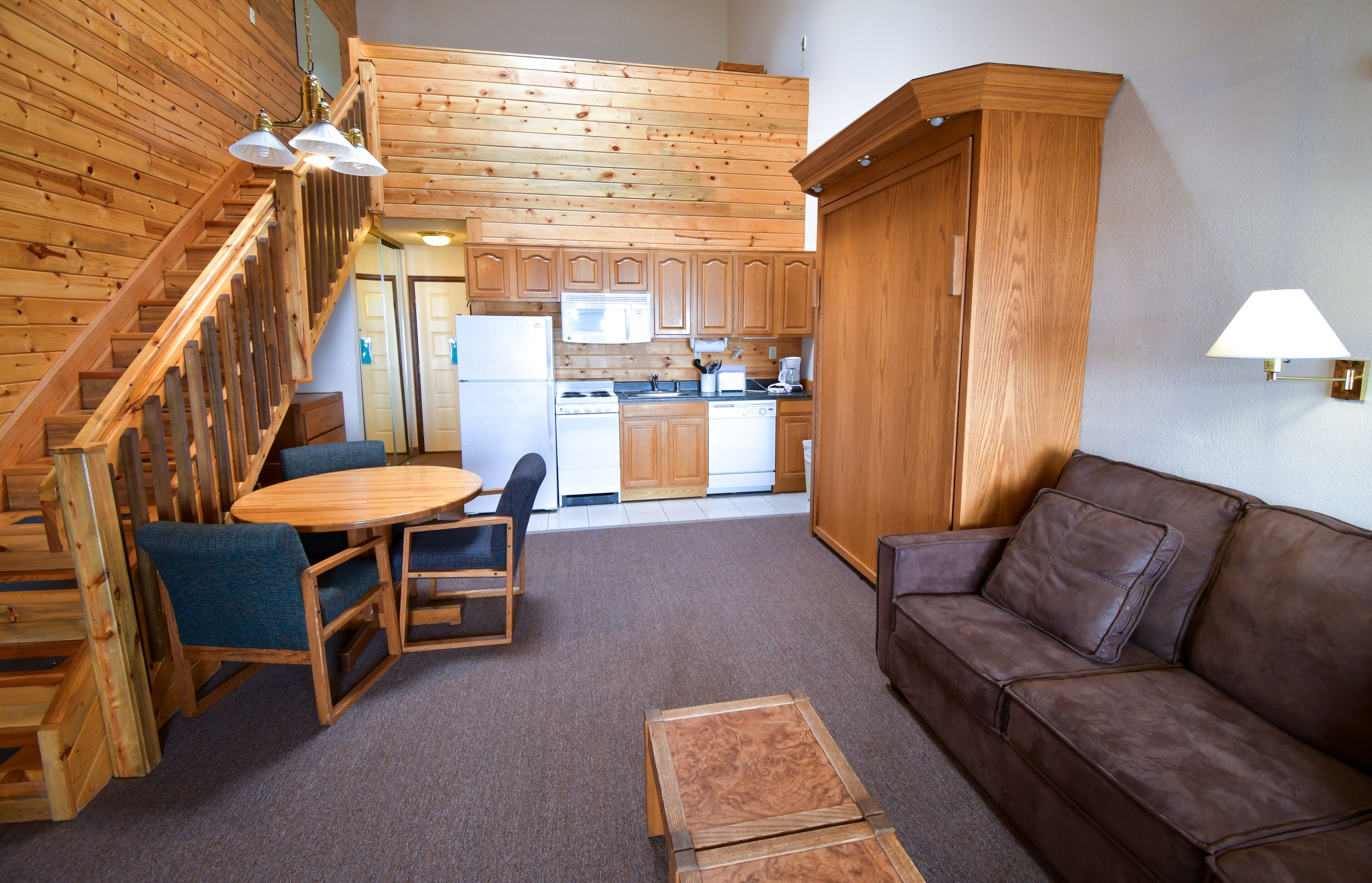 Uma cozinha compacta com armários de madeira, micro-ondas e geladeira. Há um espaço de jantar com mesa e cadeiras, e um sofá de couro marrom. Uma escada leva a um andar superior.