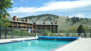 A swimming pool with a blue tarp covering part of it, surrounded by a fence. In the background, there is a building with multiple balconies and a hilly landscape.