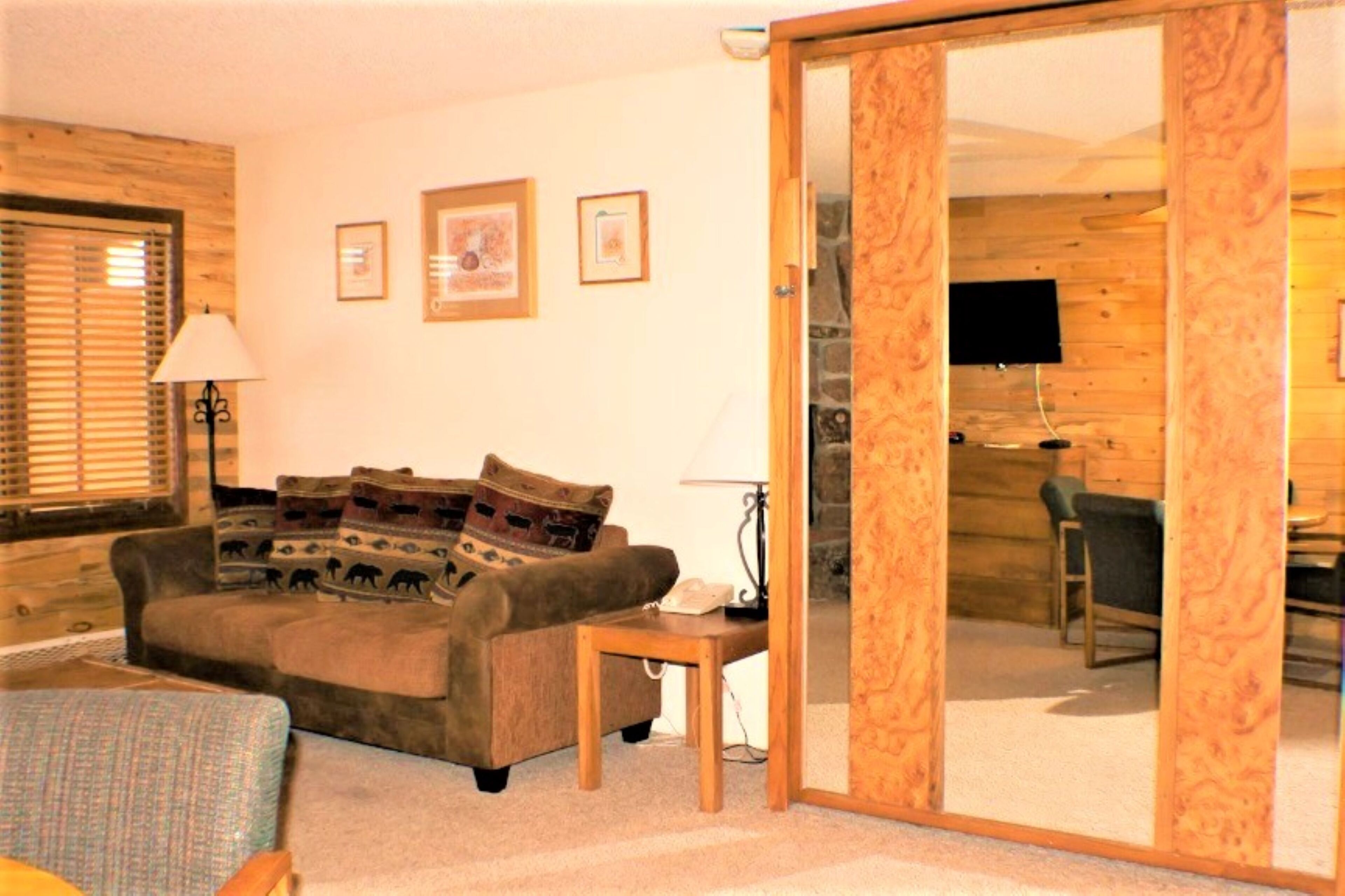A cozy living room with a sofa, a small table, and a television. There are framed pictures on the wall and a wooden door leading to another room.