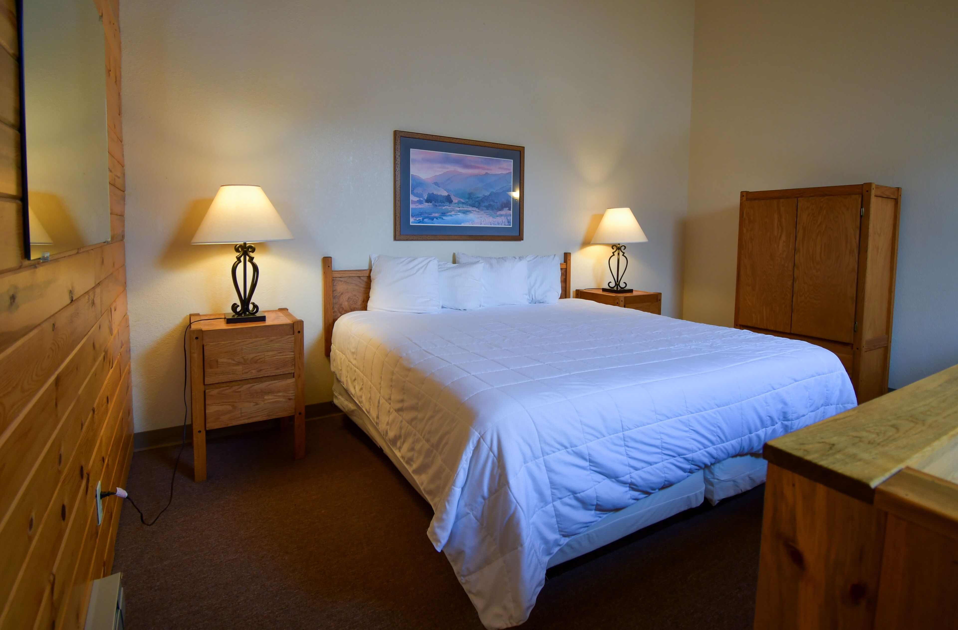 Quarto de hotel com uma cama grande, duas mesas de cabeceira com luminárias, um guarda-roupa de madeira e um quadro na parede.