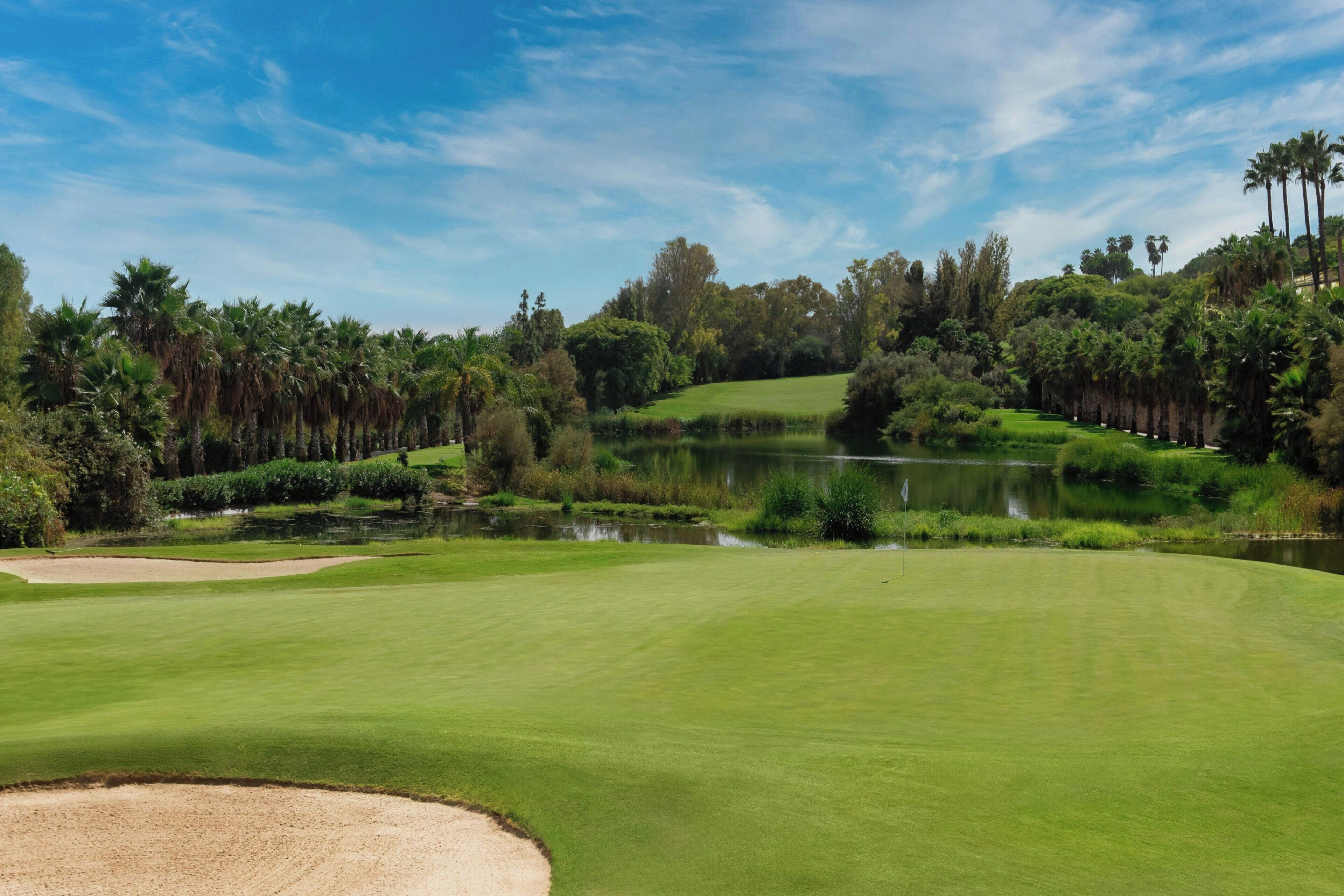 Een golfbaan met een vijver, zandbunkers en dichte bomen.
