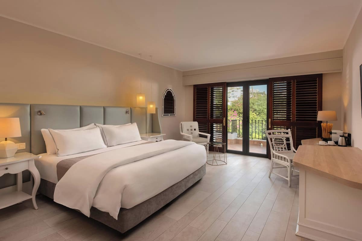 Een moderne hotelkamer met een groot bed, een bureau en een stoel. Er is een balkon met houten luiken en uitzicht op groen.