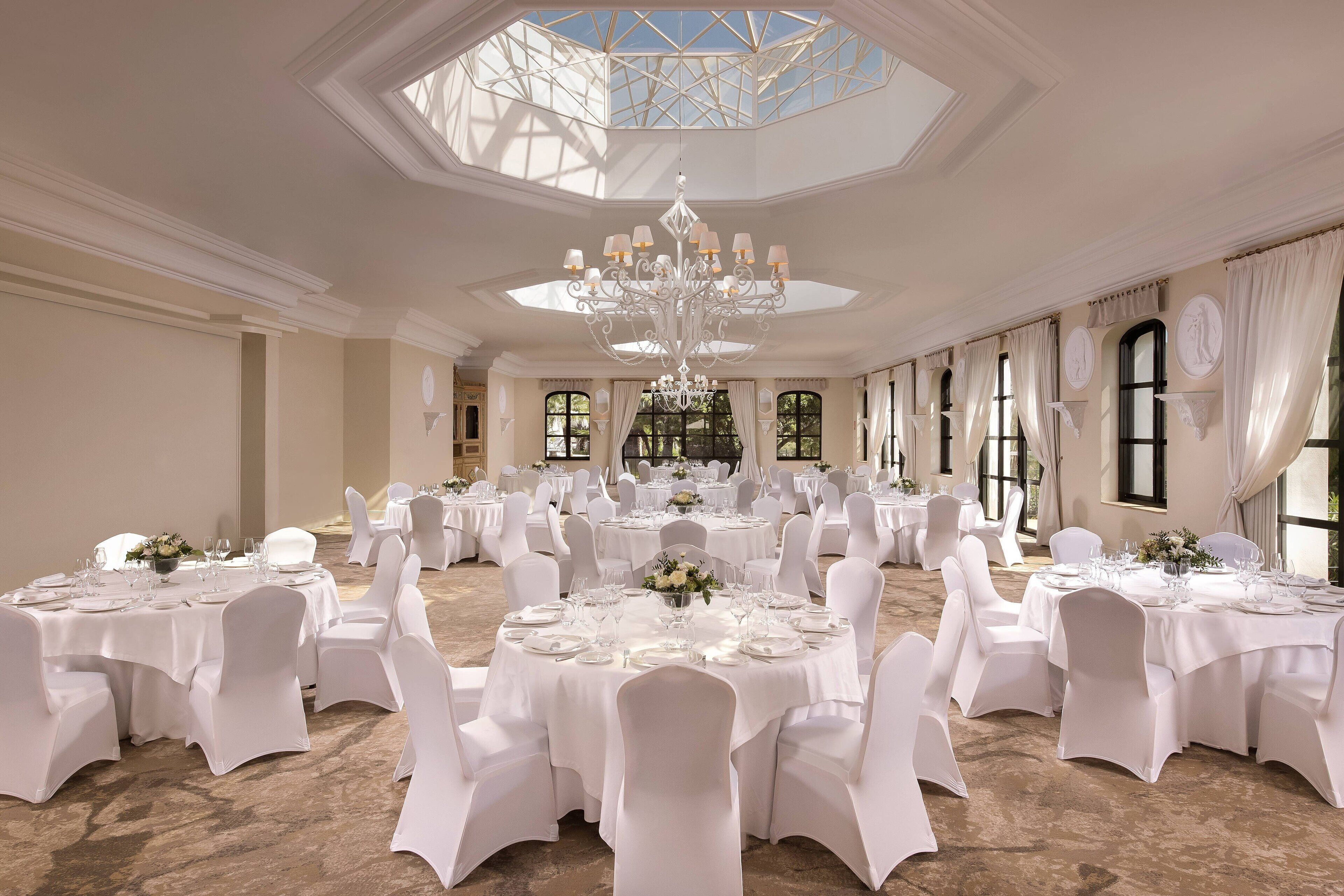 A banquet hall with round tables, white chairs, chandeliers, and large windows.