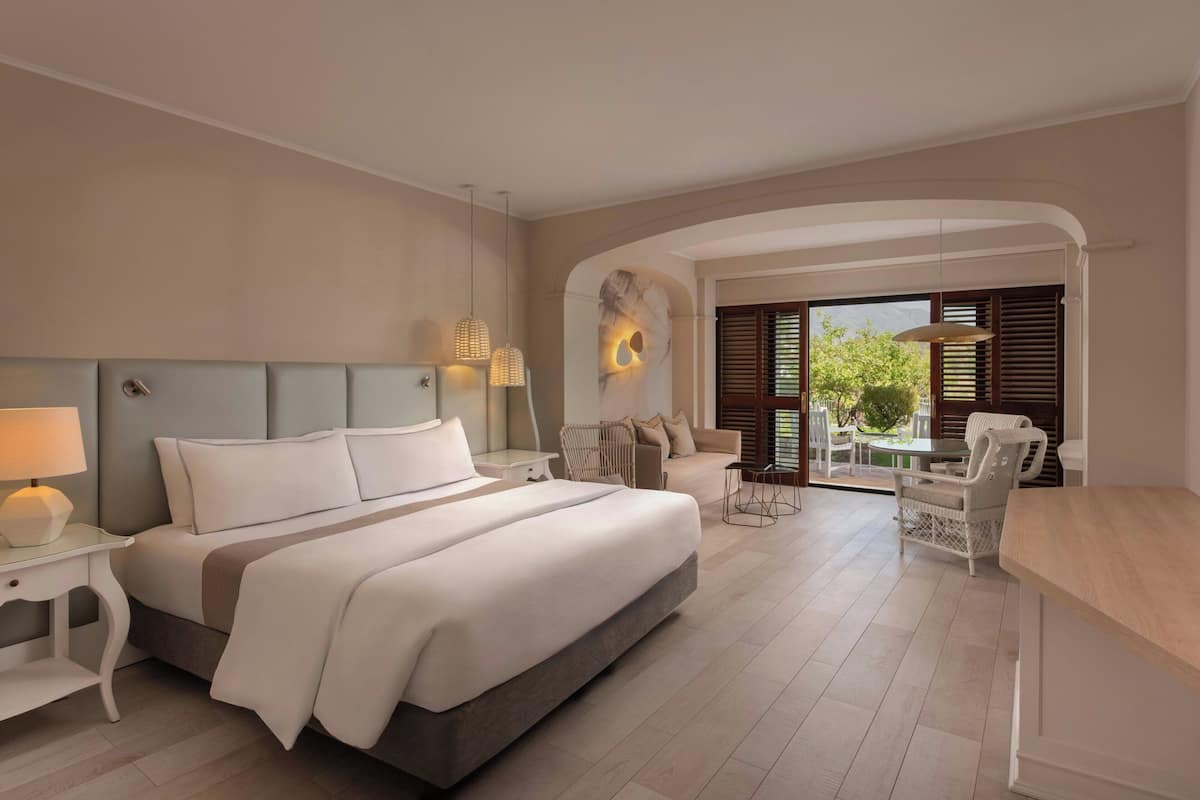 Een moderne hotelkamer met een groot bed, een zithoek met een tafel en stoelen, en uitzicht op het omliggende landschap door een schuifdeur.