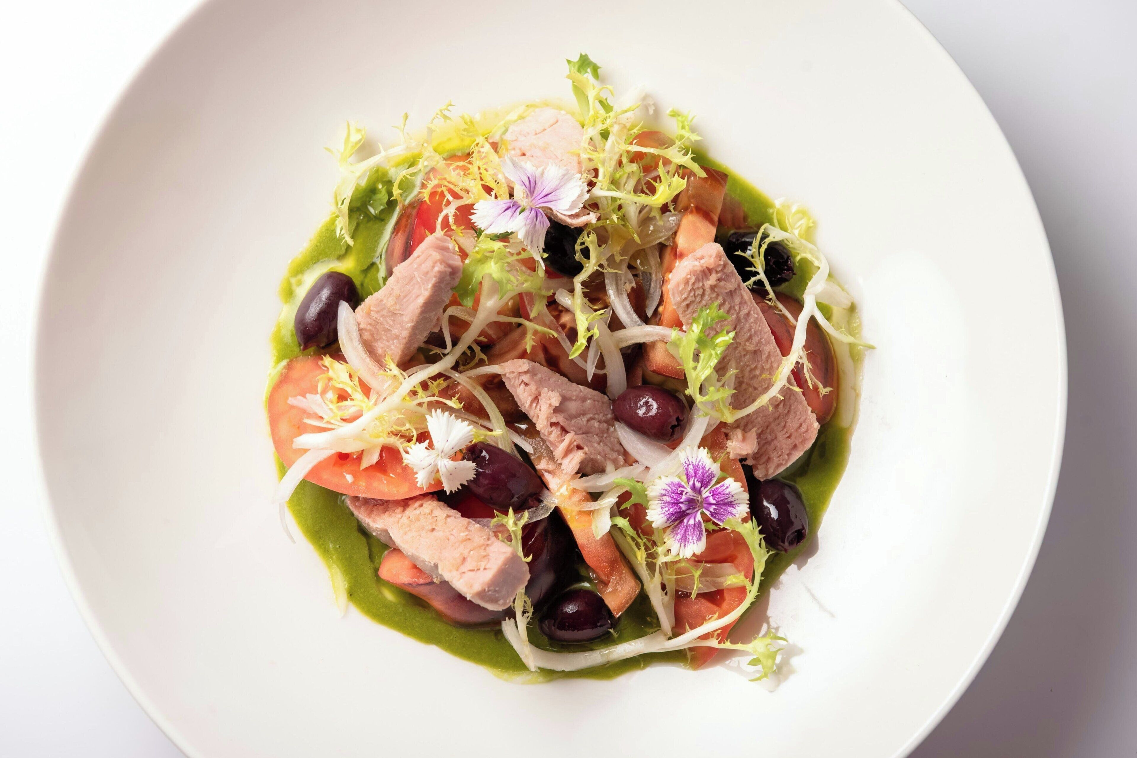 A white plate with a salad containing sliced meat, tomatoes, olives, and greens.