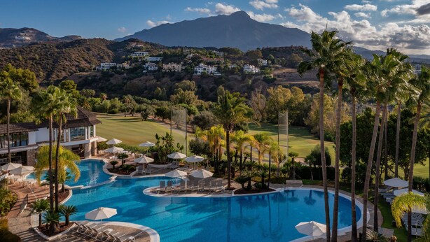 Een resort met een groot zwembad, ligbedden en parasols. Er staan palmbomen en in de achtergrond is een golfbaan en bergen te zien.