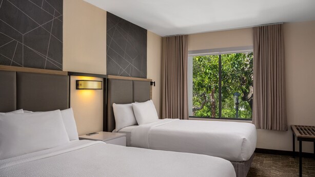 Hotelkamer met twee bedden, een nachtkastje, een lamp, een raam met gordijnen en uitzicht op groen.