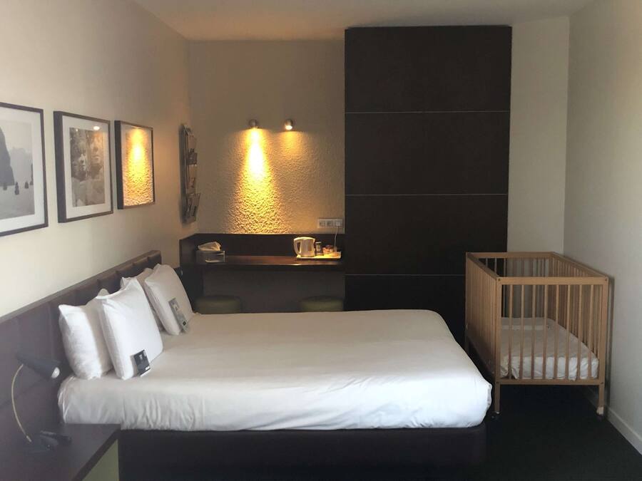 Habitación de hotel con cama, cuna de madera, escritorio con silla e iluminación en la pared.