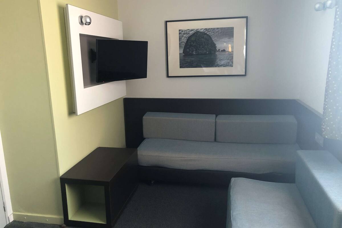 A small, modern room with a grey sofa, a flat-screen TV mounted on the wall, a framed picture above the sofa, and a small table beside the sofa.