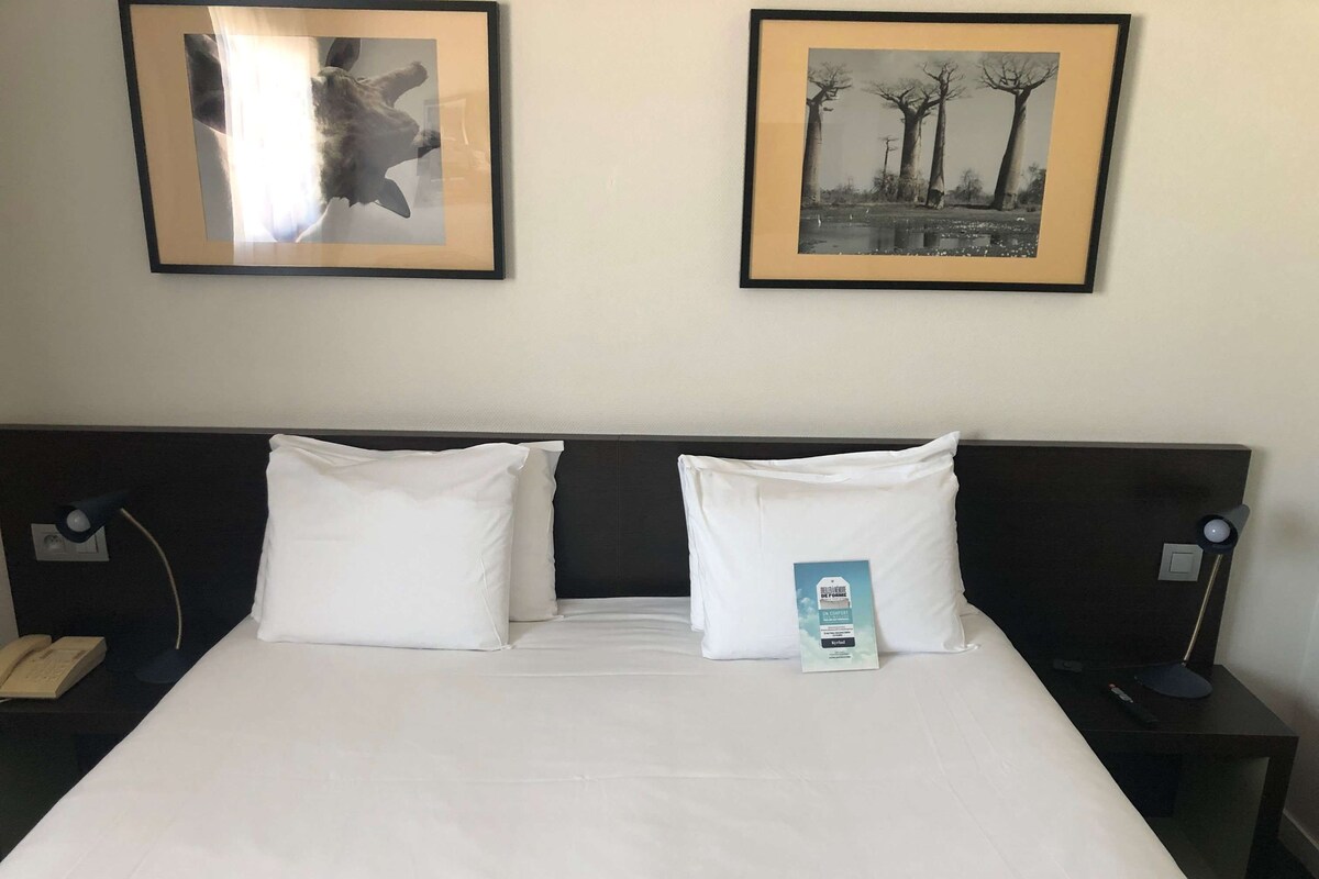 Een hotelkamer met een groot bed, twee witte kussens, een nachtkastje met een lamp en twee ingelijste foto's aan de muur.
