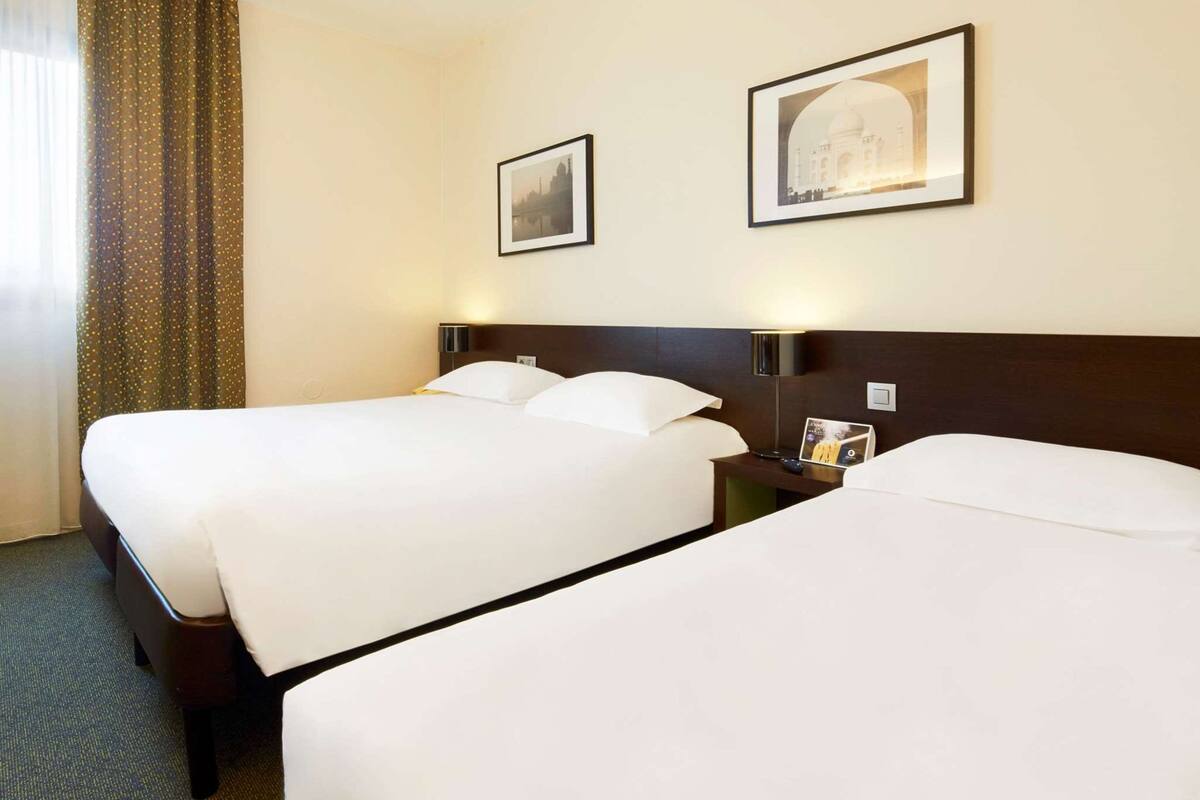 A hotel room with two beds, a nightstand with a lamp, and two framed pictures on the wall.
