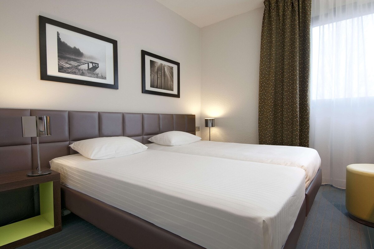 Une chambre d’hôtel avec deux lits, une table de chevet, une lampe et deux tableaux encadrés au mur.