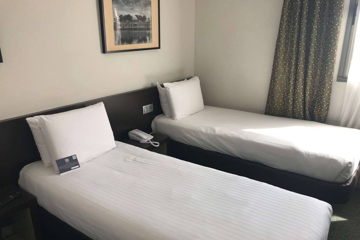 A hotel room with two single beds, a bedside phone, a framed picture on the wall, and a window with curtains.
