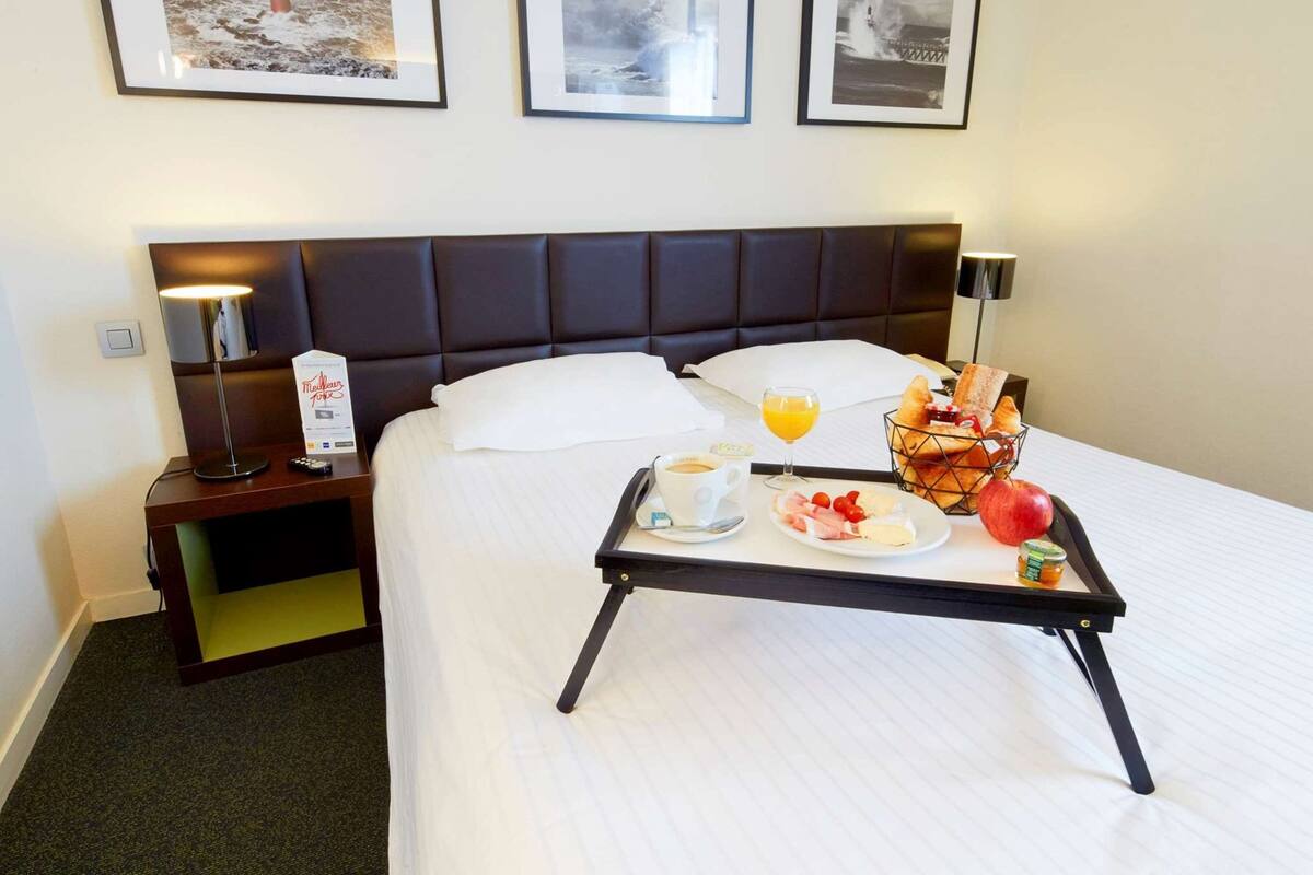 A hotel room with a neatly made bed, a bedside table with a lamp, a tray with breakfast items, and three framed pictures on the wall.