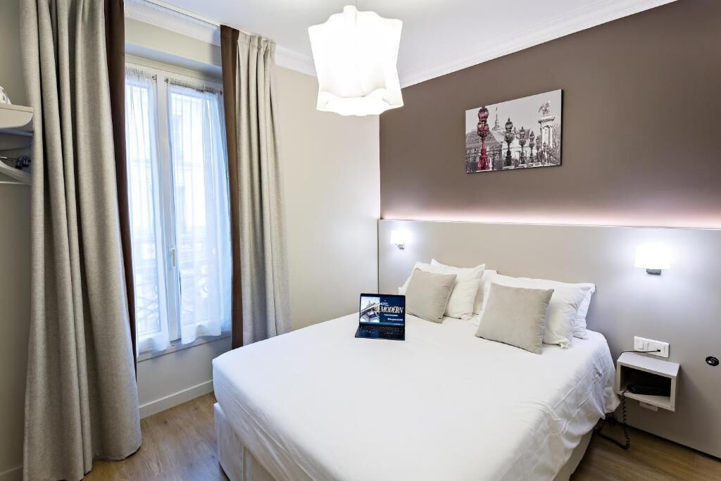 Un cuarto de hotel con una cama grande, una mesita de noche, una lámpara, un cuadro en la pared y una ventana con cortinas.