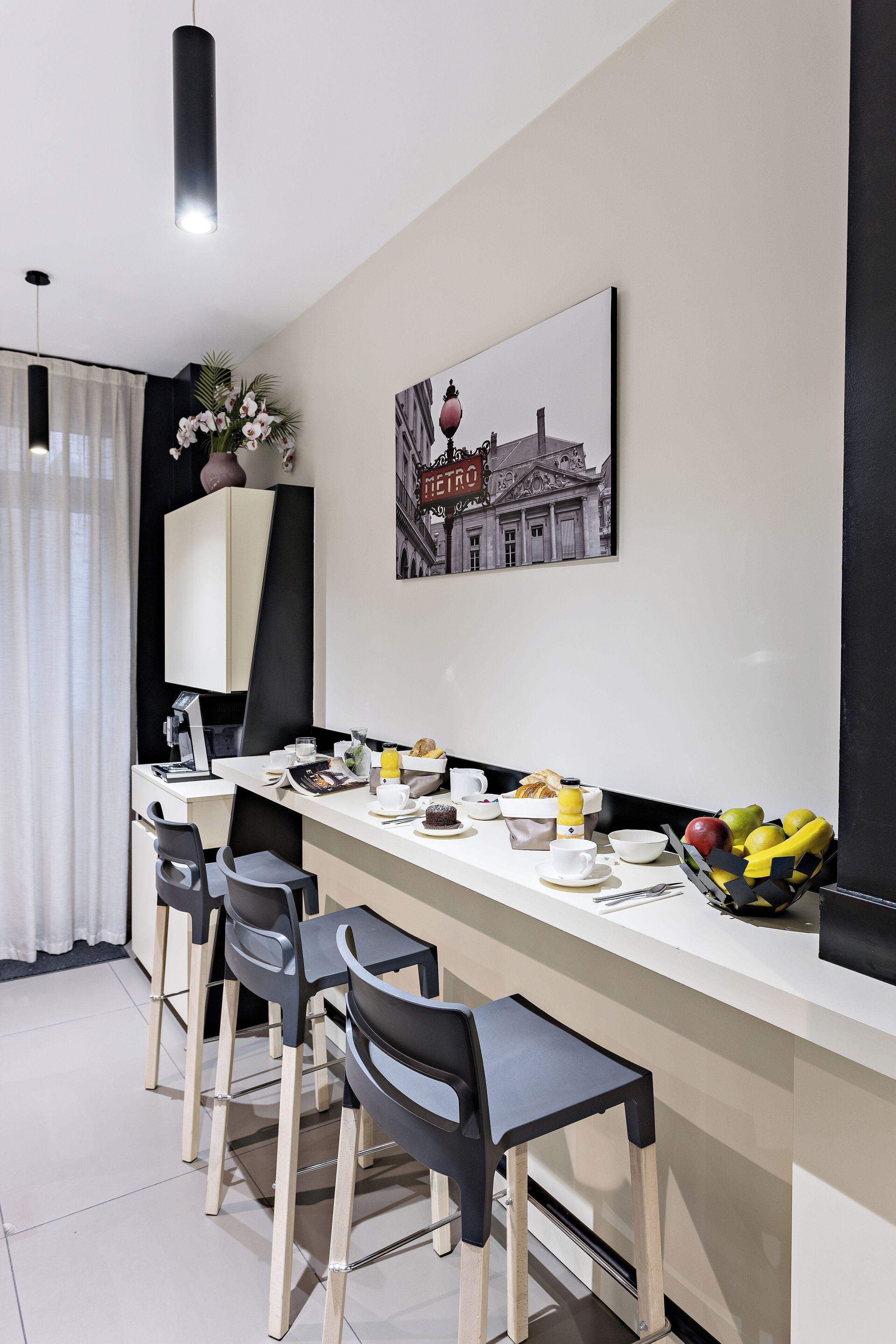 A modern kitchen with a long counter, bar stools, and a dining area. There is a framed picture on the wall and a vase with flowers on the counter.
