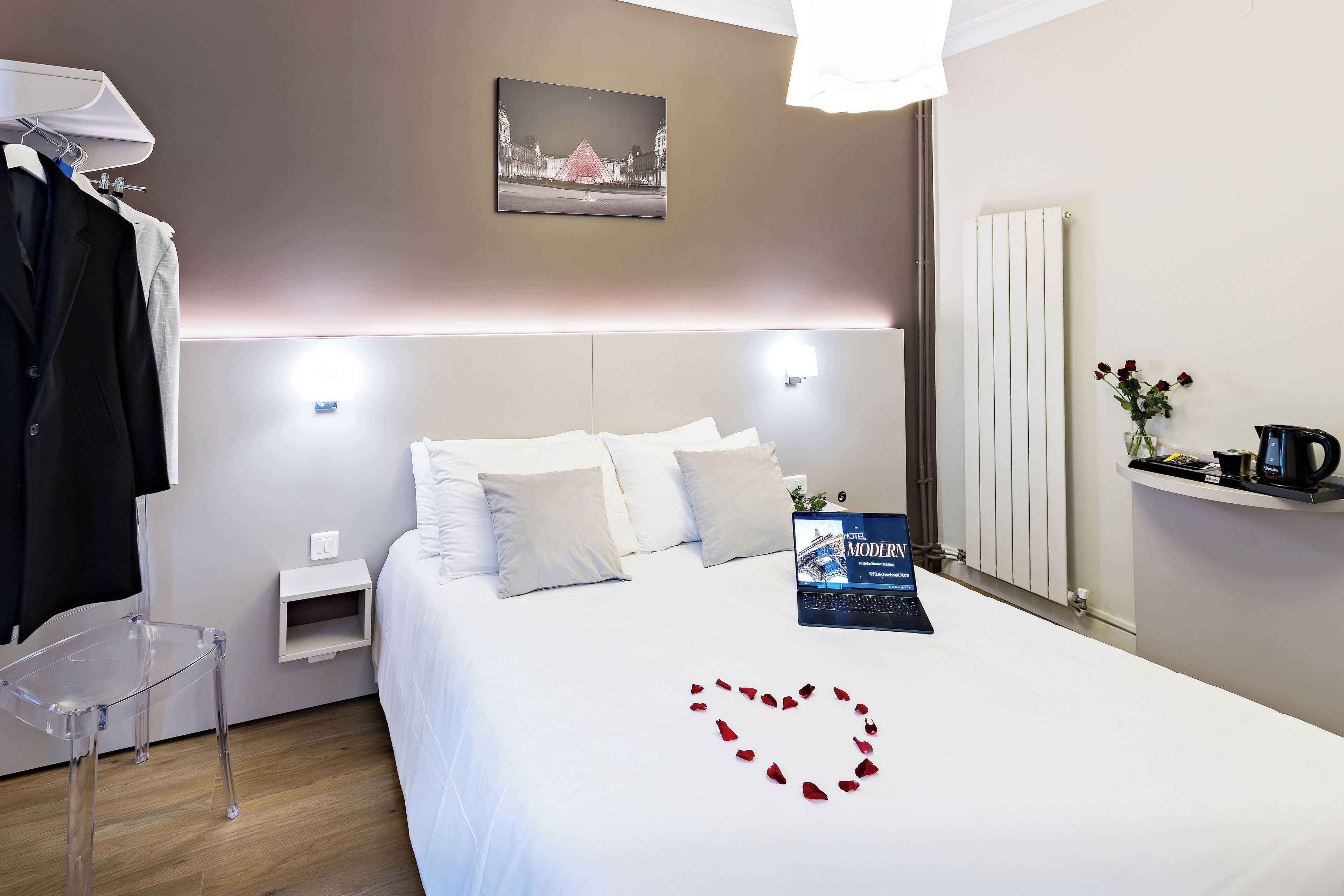 A hotel room with a bed, a laptop, a clear chair, a bedside table, a radiator, and a wall-mounted picture.