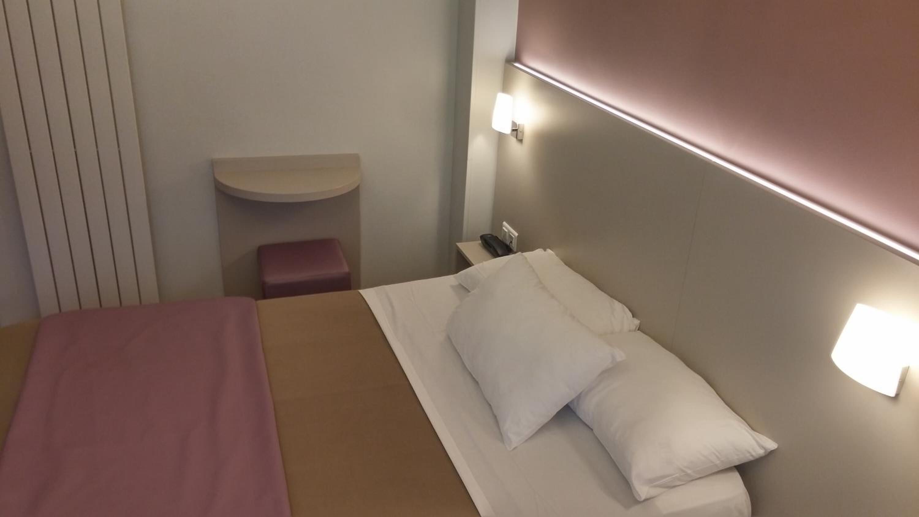 Hotellihuone, jossa on sänky, yöpöytä, pieni sivupöytä ja seinään kiinnitetty valaisin.