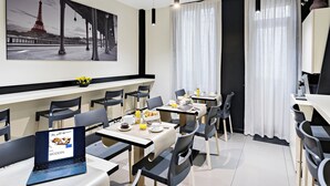 Habitació d'hotel moderna amb zona de menjador, un ordinador portàtil sobre la taula, articles per l'esmorzar i una obra d'art gran amb la Torre Eiffel a la paret.