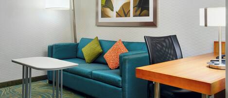 A hotel room with a teal sofa, a small white table, a wooden desk, and a framed artwork on the wall.