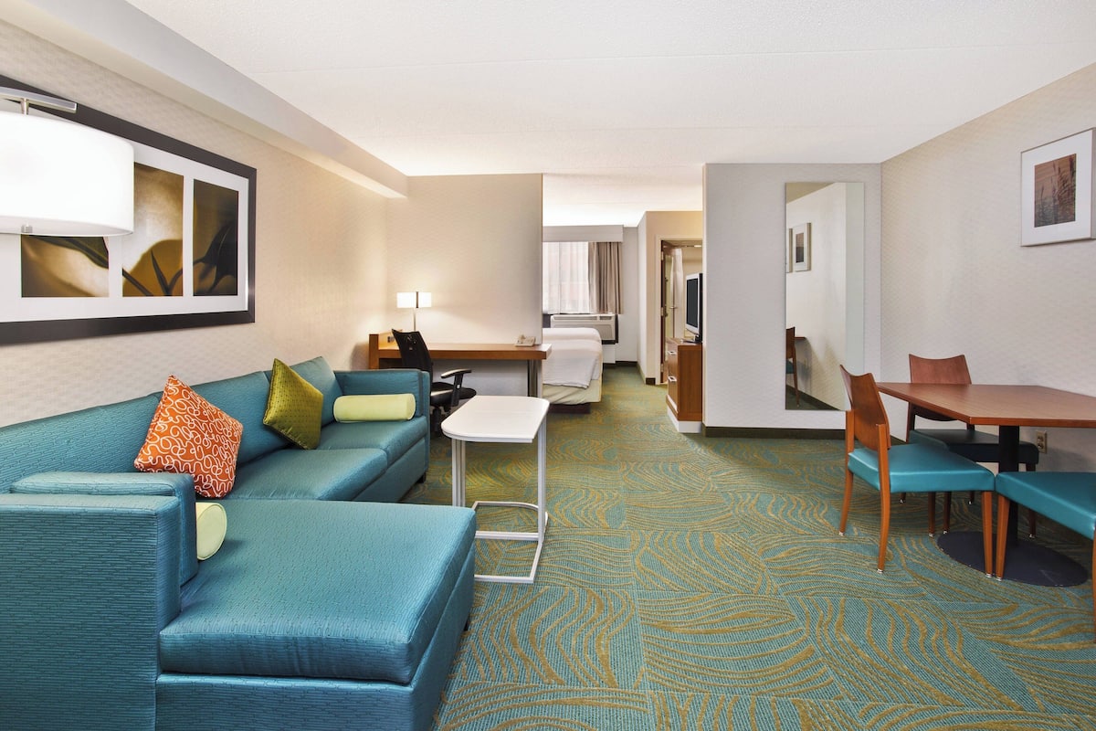 A hotel room with a blue sofa, a small table, a desk, and a dining area with chairs.