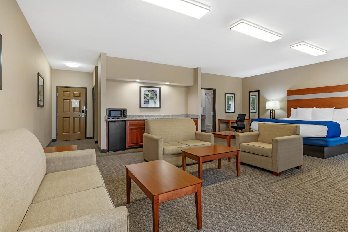 A hotel room with a sofa, two armchairs, a coffee table, a bed with blue bedding, a desk, and a kitchenette.