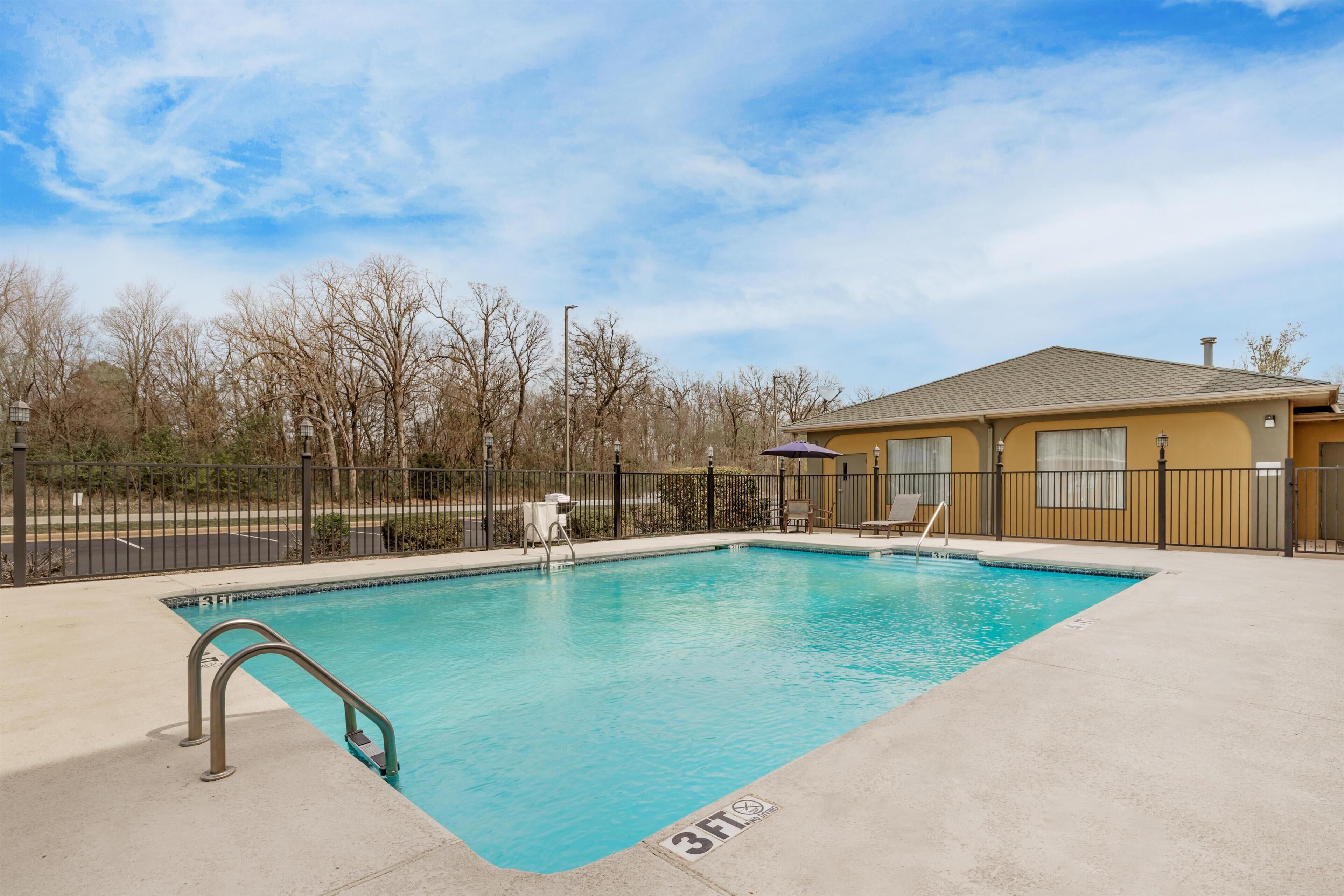 A rectangular swimming pool with a metal ladder on one side, a small deck area with chairs, and a building with a brown roof in the background.