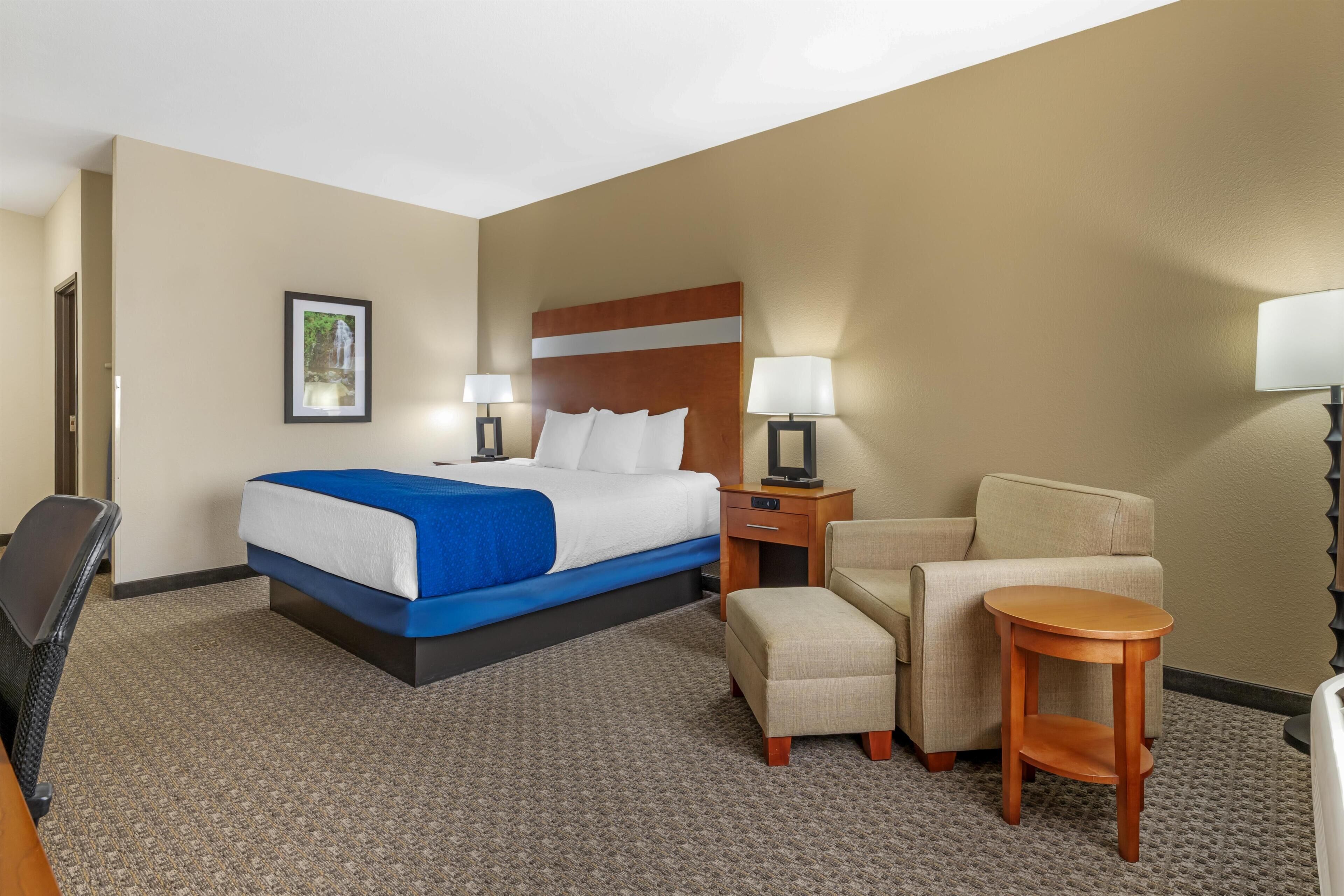 A hotel room with a bed, a desk, a chair, a small table, and a lamp.