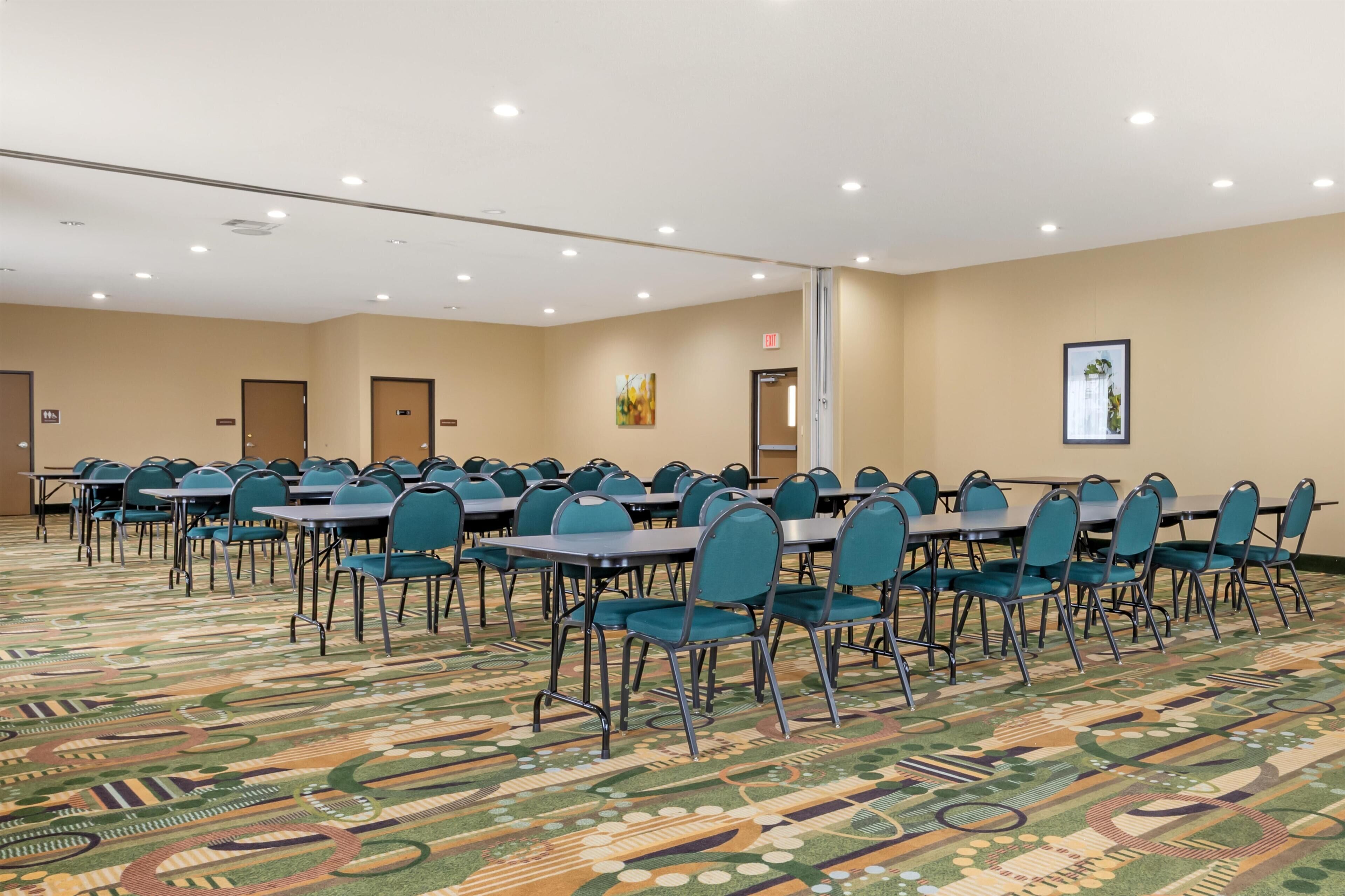 Un salón de conferencias con mesas y sillas dispuestas en filas, suelo alfombrado e iluminación en el techo.