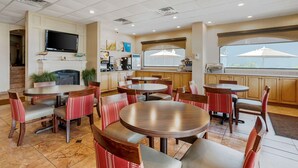 A restaurant with multiple round tables and chairs, a flat-screen TV mounted on the wall, and a window with an umbrella. - Comfort Inn Ocean City Boardwalk (Ocean City)
