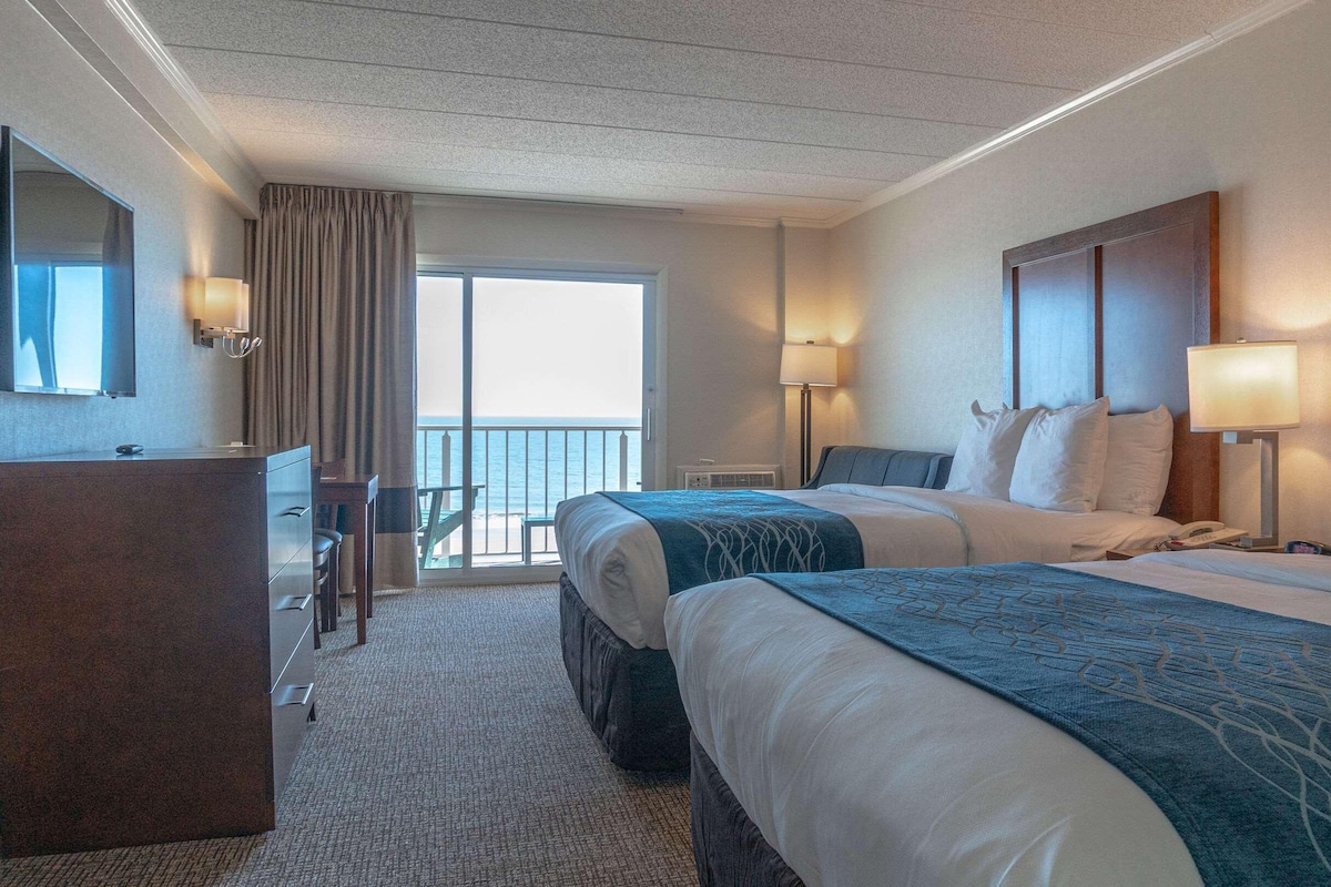 A hotel room with two beds, a desk, a chair, and a balcony with a view of the sea.