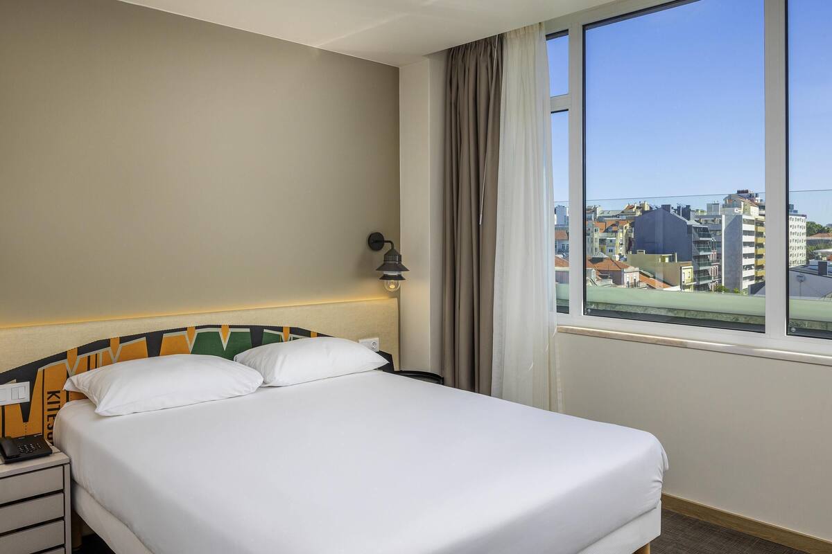 Une chambre d’hôtel comprenant un lit, un téléphone sur la table de chevet, une lampe, une fenêtre avec des rideaux et une vue sur les bâtiments.