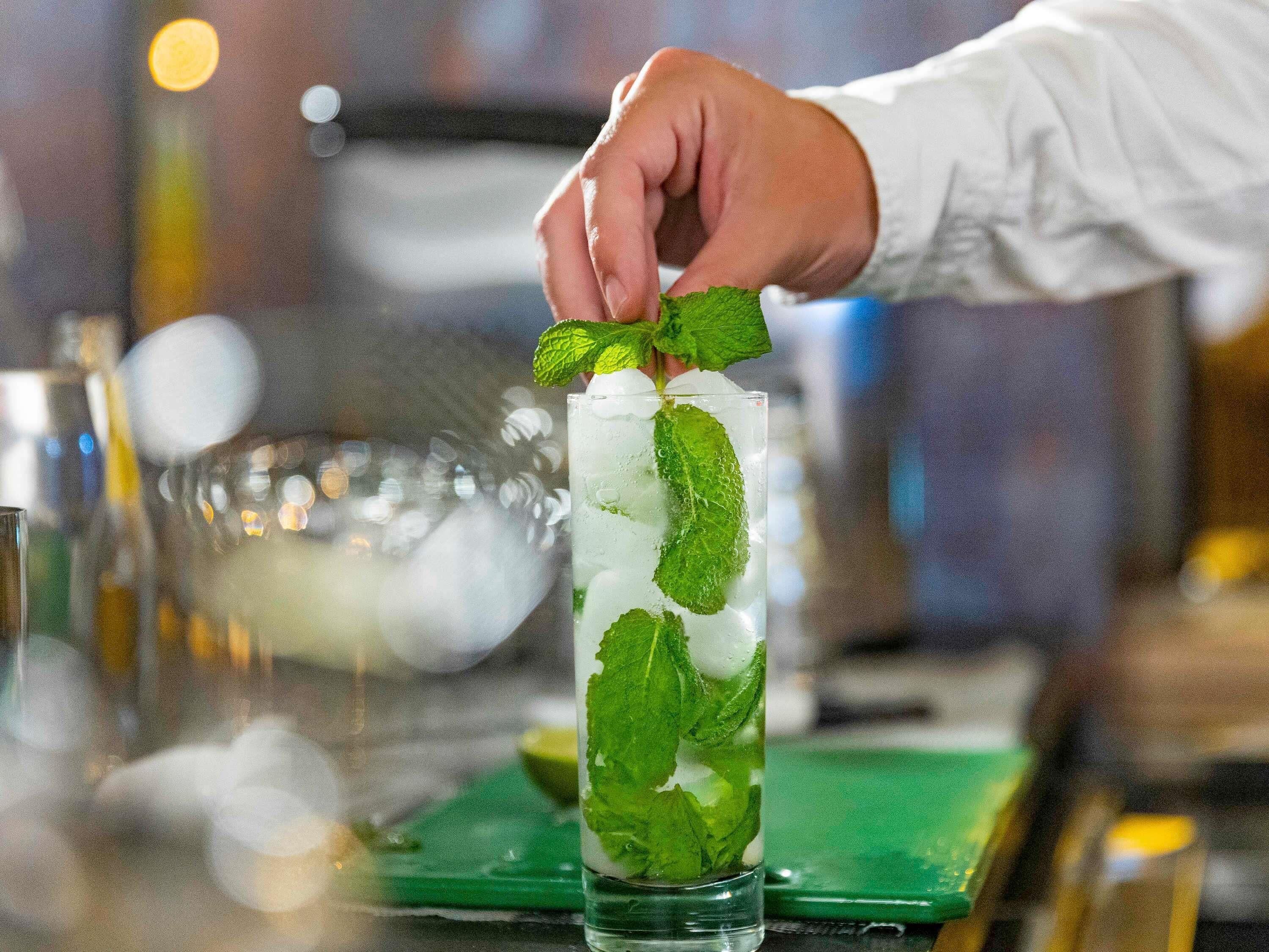 Una mano agregando hojas de menta a un vaso alto lleno de hielo y un líquido transparente, probablemente un cóctel.