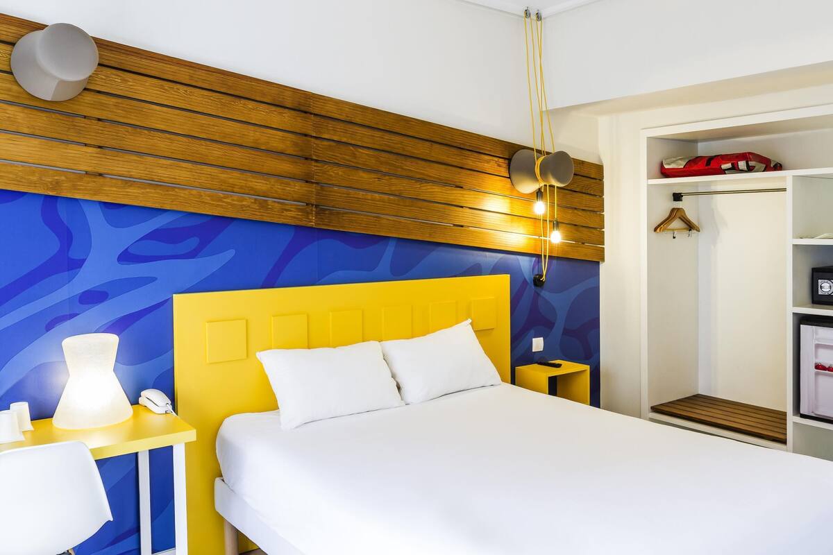 Une chambre d’hôtel avec un plafond en lambris de bois, une tête de lit jaune, un lit blanc, une table de chevet avec une lampe et une armoire avec une tringle pour suspendre les vêtements.