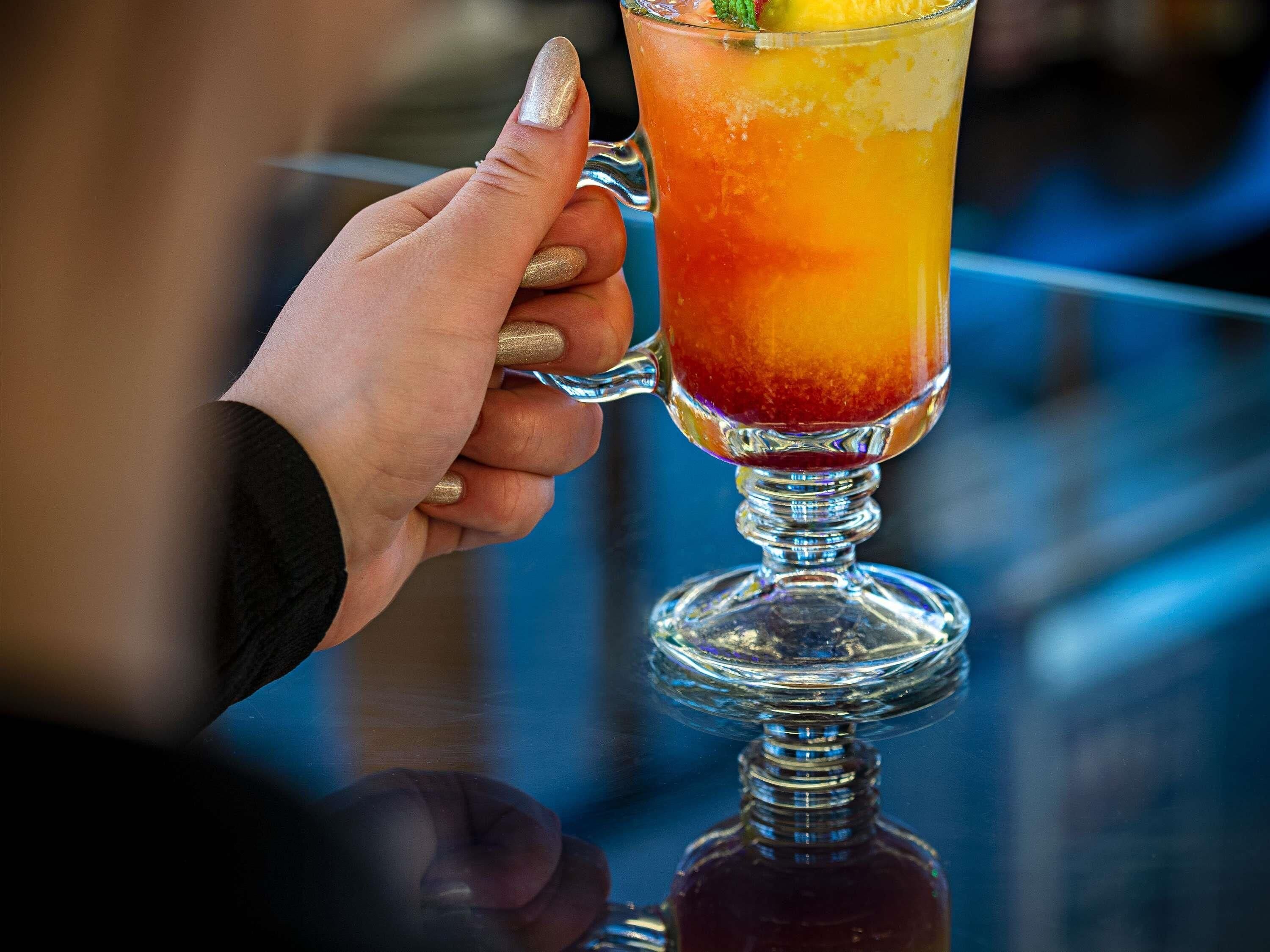 A hand holding a tall glass with a gradient drink, possibly a cocktail, with a lime wedge on the rim.
