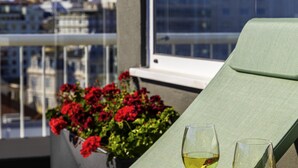 Un balcon avec une chaise verte et deux verres de vin blanc, donnant sur un paysage urbain avec des bâtiments et des fleurs rouges en arrière-plan.