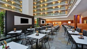 Un vaste espace intérieur aménagé pour les repas, avec de nombreuses tables et chaises, une plante centrale et un design moderne au niveau du plafond.