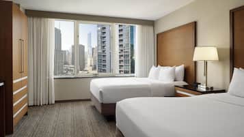 Habitación de hotel con dos camas, un ventanal grande con vistas a la ciudad, un armario de madera y una mesita de noche con lámpara.