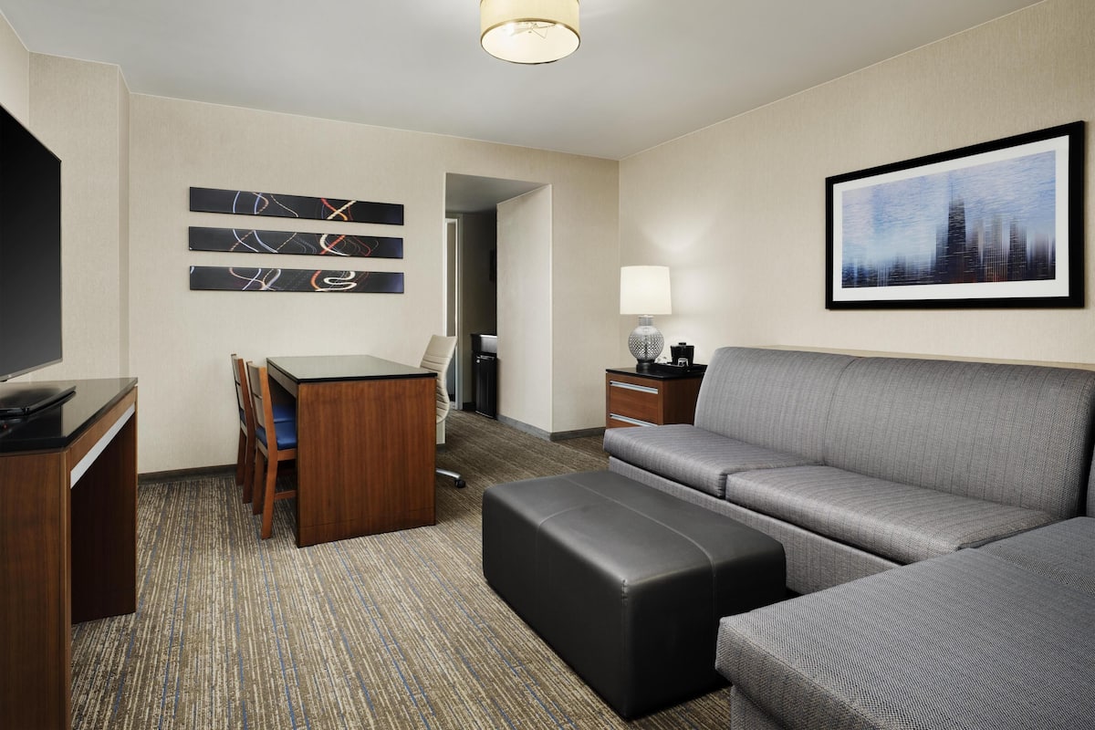 A hotel room with a sofa, ottoman, desk, chair, TV, artwork, lamp, and a framed picture on the wall.