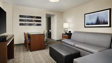 A hotel room with a flat-screen TV, a wooden desk, a grey sofa, a black ottoman, a framed picture on the wall, and a lamp on a side table.