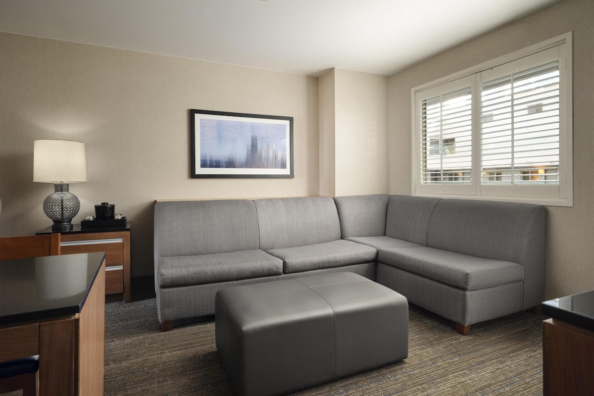 A modern hotel room with a gray sectional sofa, a small ottoman, a desk with a lamp, and a framed picture on the wall.