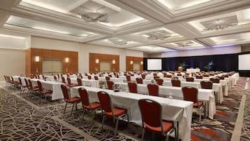 A conference room with rows of tables and chairs, a stage with a screen, and a patterned carpet.