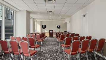 Un salón de conferencias con filas de sillas rojas, una alfombra estampada y un ventanal grande en un lateral.
