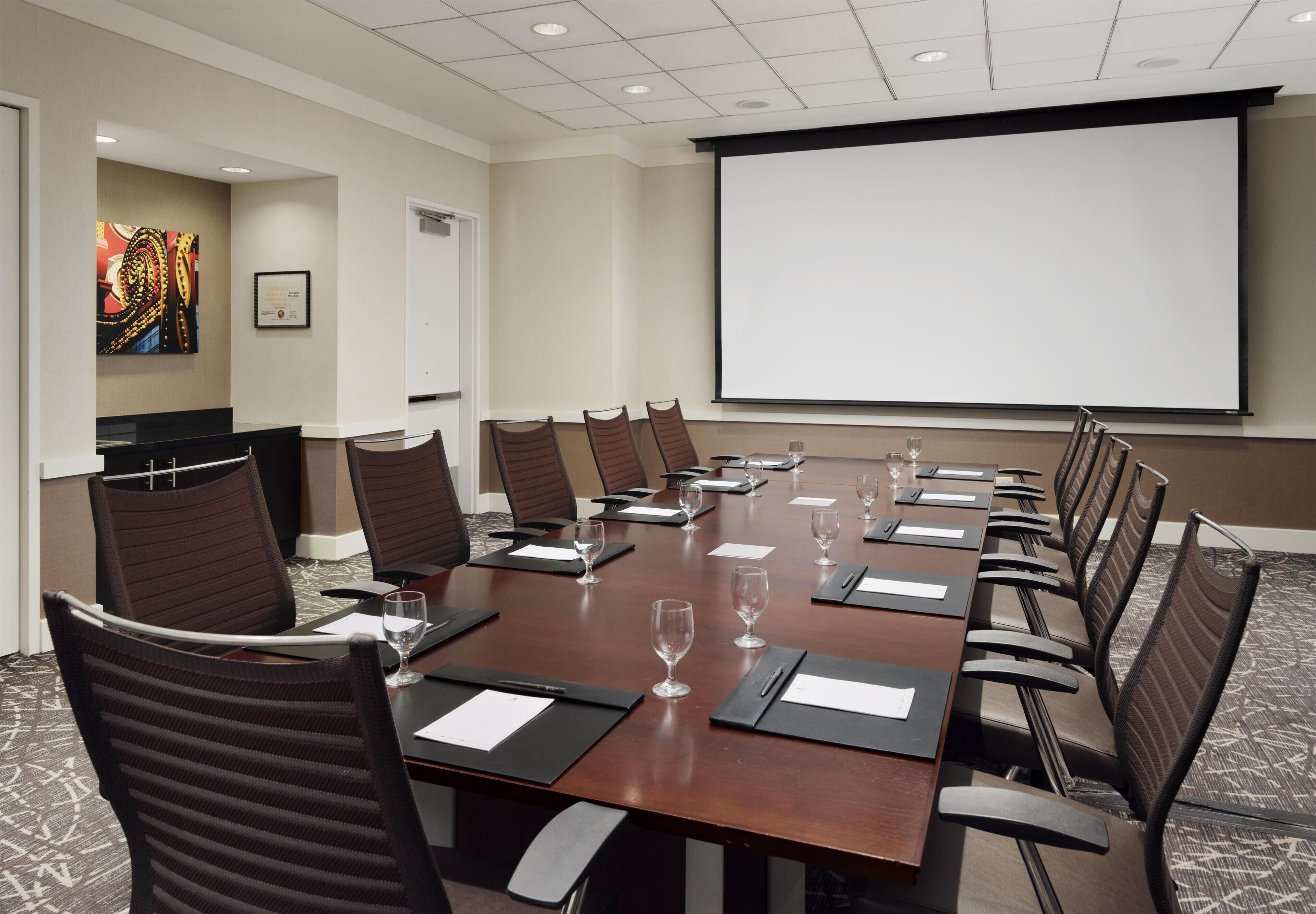 A conference room with a long table, chairs, a projector screen, and a carpeted floor.