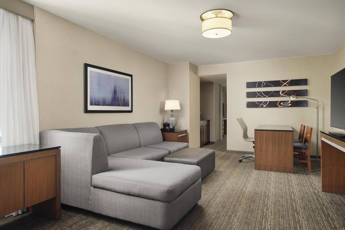 A hotel room with a gray sectional sofa, a desk, a chair, a lamp, and artwork on the walls.