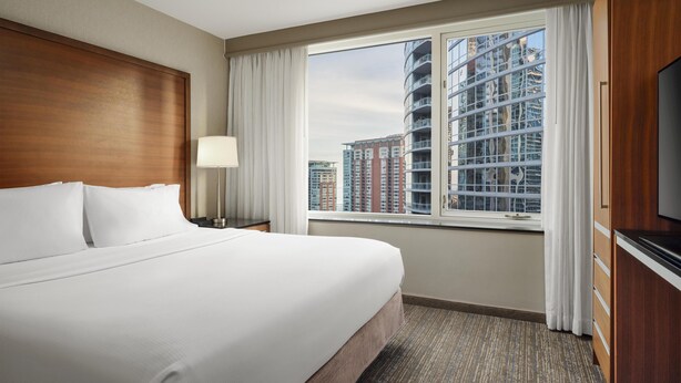 Habitación de hotel con una cama grande, un televisor, una ventana con cortinas y vistas a edificios altos.