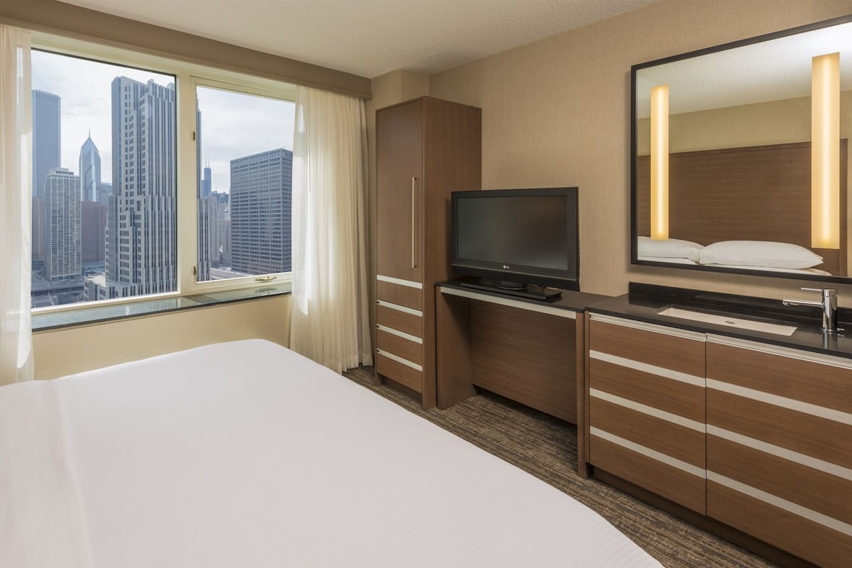 Habitación de hotel con cama, escritorio, televisor, espejo y ventana con vista a la ciudad.